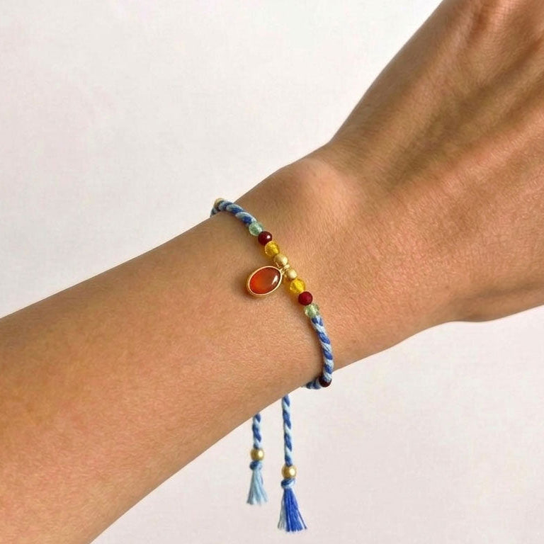 A close-up of a person's wrist wearing a colorful braided cord bracelet with a small oval orange stone and tassel details.
