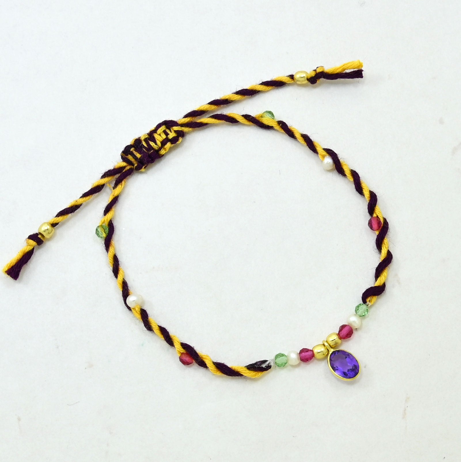 A braided cord bracelet with a purple amethyst gemstone charm, featuring yellow and dark purple twisted cords, small colorful beads, and gold-toned spacer details.