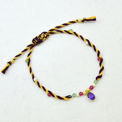 A braided cord bracelet with a purple amethyst gemstone charm, featuring yellow and dark purple twisted cords, small colorful beads, and gold-toned spacer details.