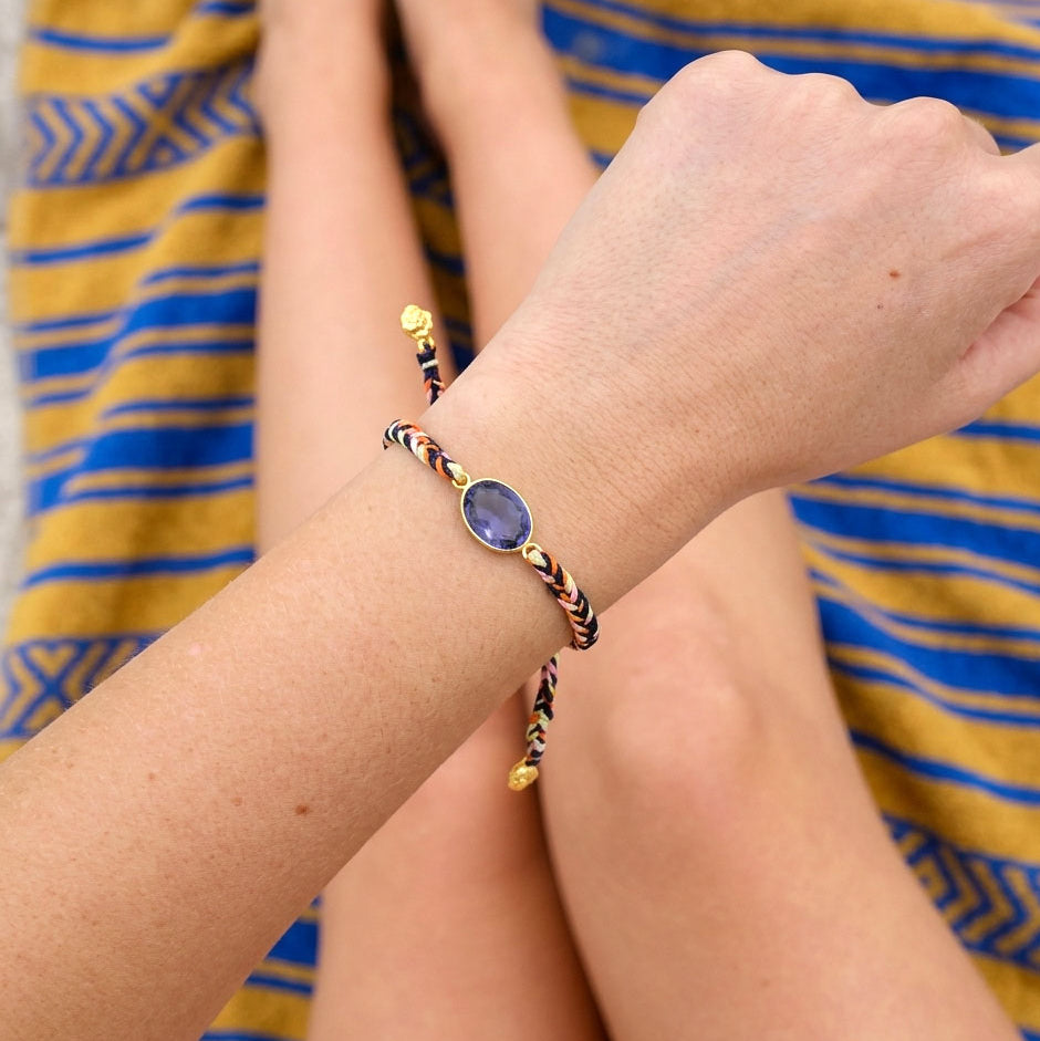 A close-up of a person's wrist wearing a braided cord bracelet with a central oval blue gemstone and small gold-toned accents.