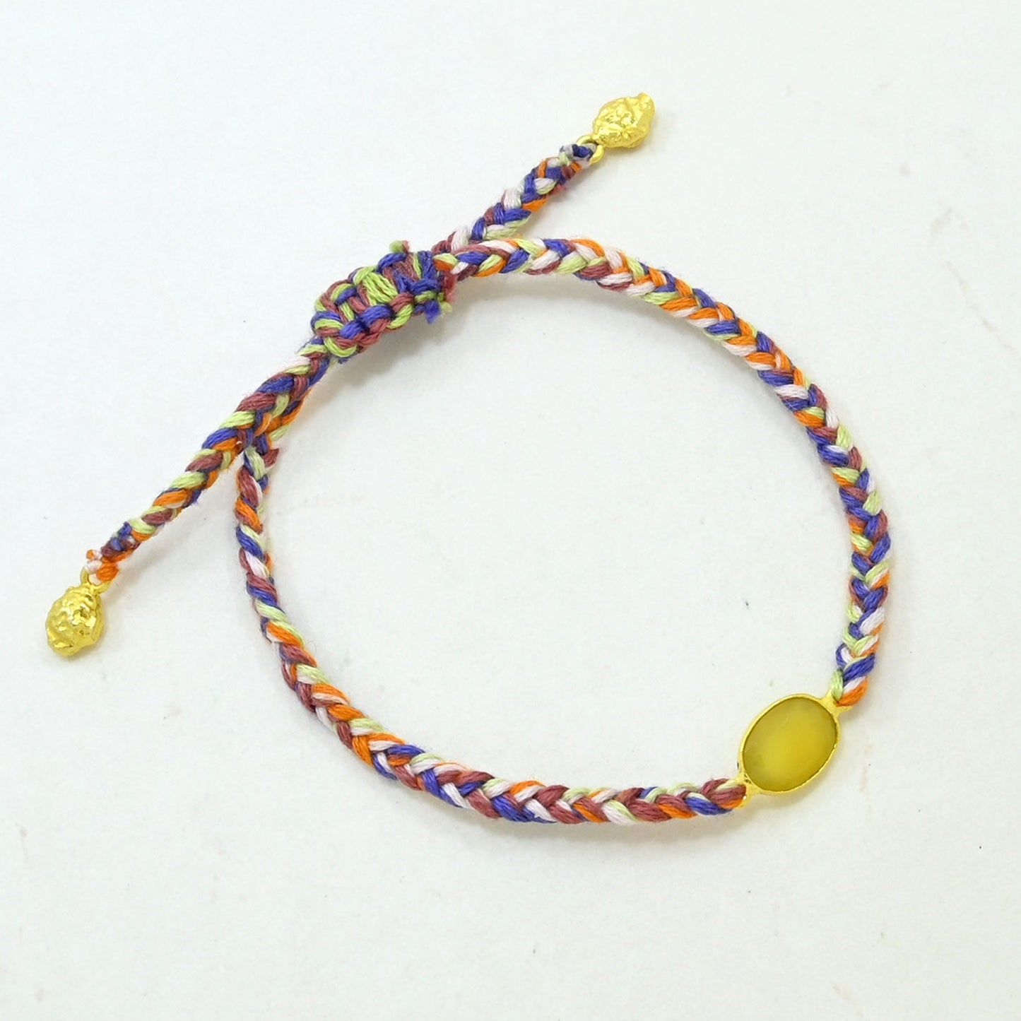 Hand braided boho bracelet with a multicolor pattern of purple, orange, white, and blue, featuring a smooth oval yellow-green bead and textured gold-tone end caps.