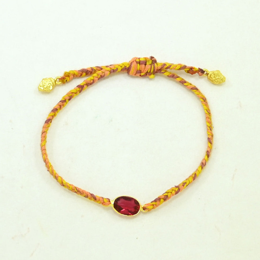 A braided cord bracelet in yellow and orange tones with a central oval red gemstone in a gold setting, plus small gold beads at the ends, shown on a plain light background.