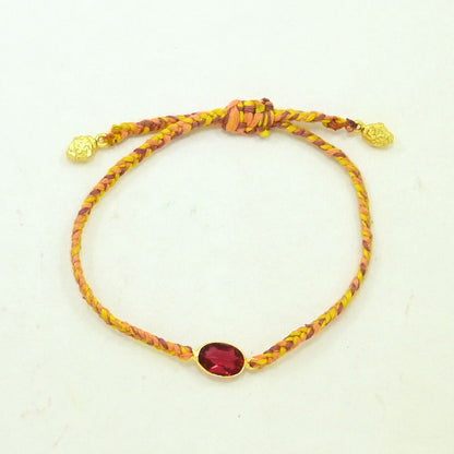 A braided cord bracelet in yellow and orange tones with a central oval red gemstone in a gold setting, plus small gold beads at the ends, shown on a plain light background.