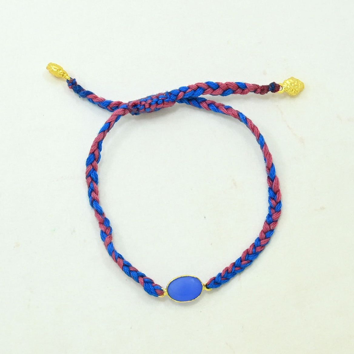 A braided cord bracelet with blue and red strands, featuring a central oval blue stone and gold-tone metal accents, including a small bead and adjustable sliding knot ends.
