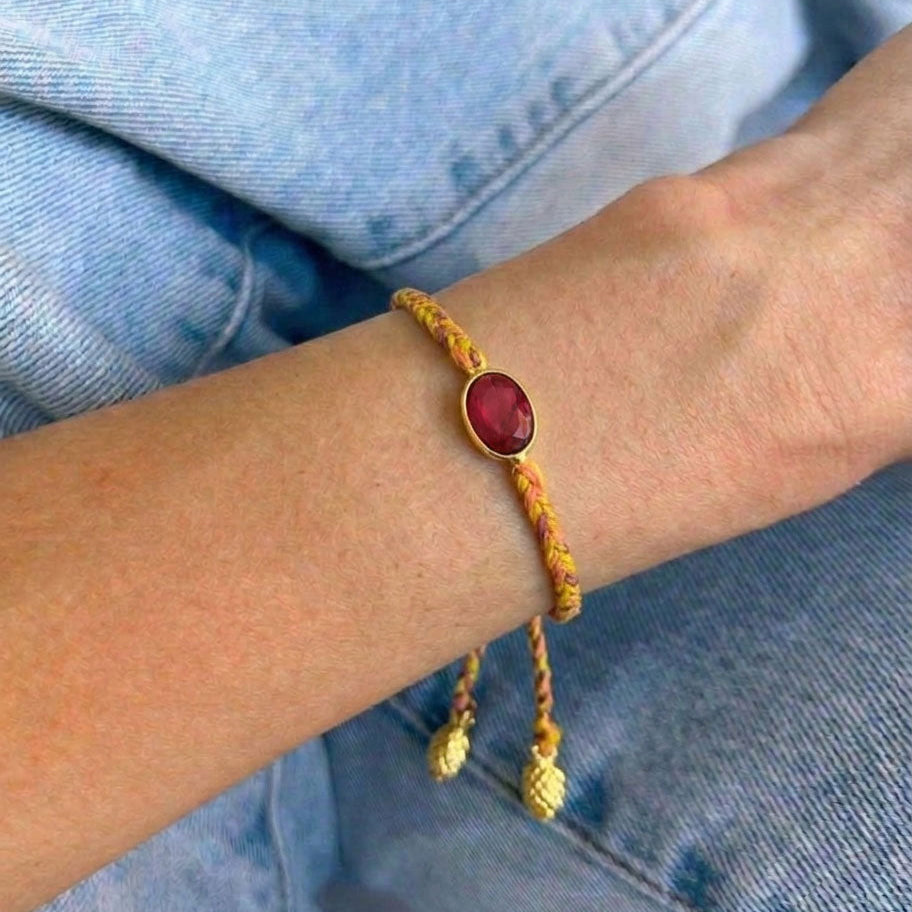 Yellow orange braided cord bracelet with a central oval pink gemstone and small dangling gold beads, set against light blue denim clothing.